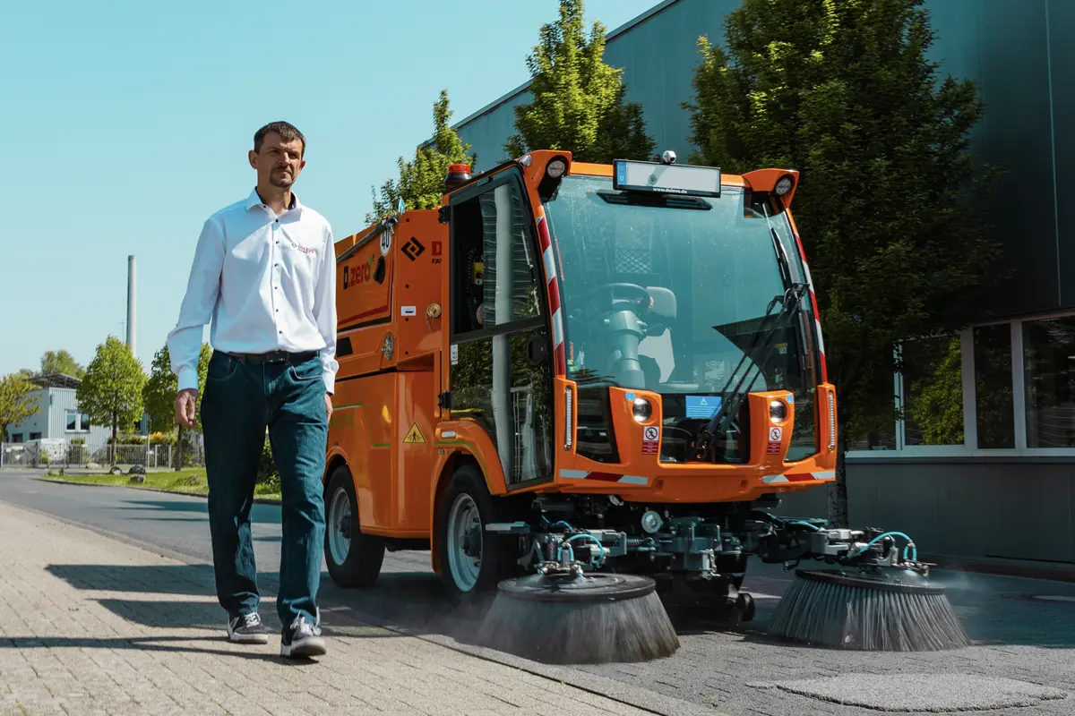 a person walking on the sidewalk while a road sweeper moves past him