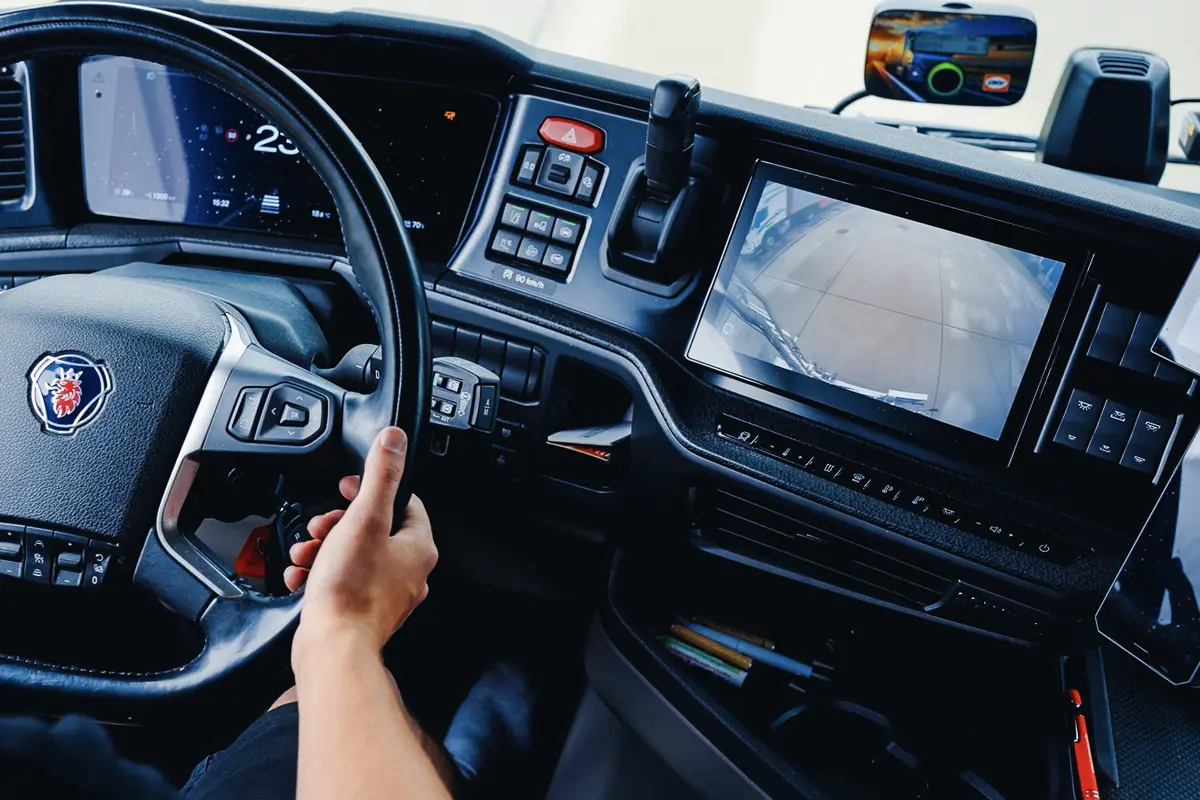 Intérieur de la cabine d'un camion Scania : le conducteur a les mains sur le volant et un écran affiche une vue de l'arrière du camion